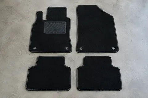 Quatre tapis sur mesure en moquette noire à surpiqûre blanche, dont un avec talonnette, posés à plat sur un sol de garage gris clair